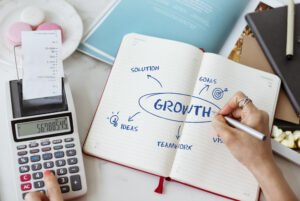 A notebook open with the word growth in capital letters. A right hand holding a pen is poised above the notebook and a left hand is typing on a calculator.