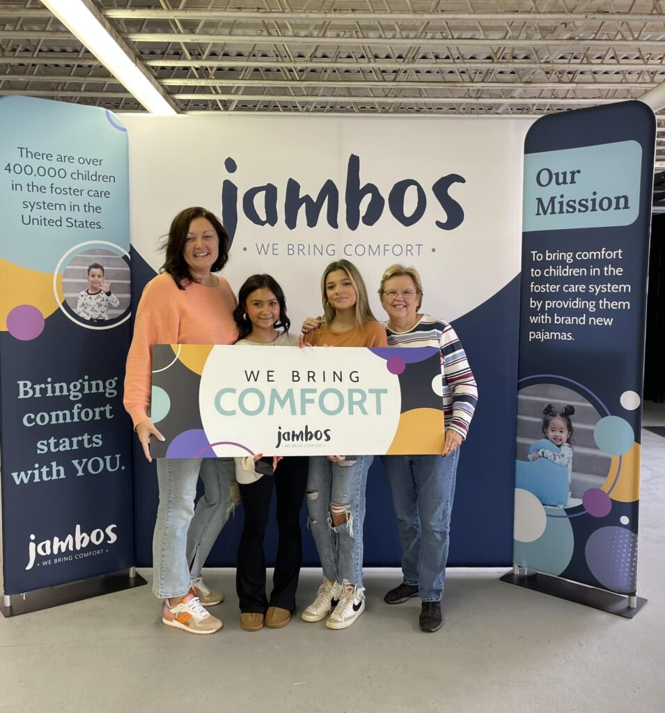 Four women stand holding a sign that says "We Bring Comfort." Behind them is a large trifold backdrop that says jambos.