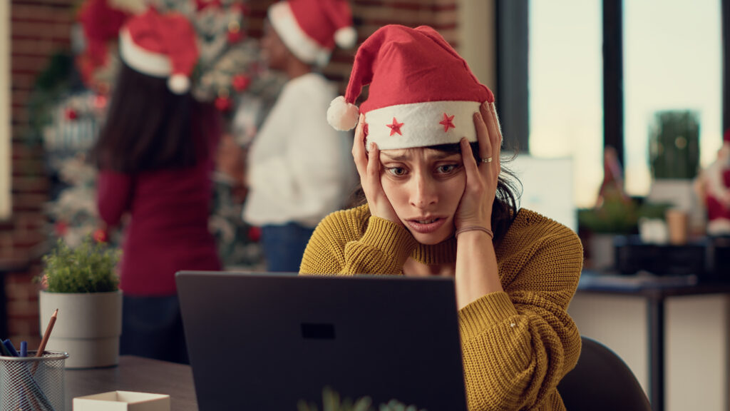 Woman in santa hat sits with her head in her hand, looking at laptop, while colleagues decorate a tree behind her.
