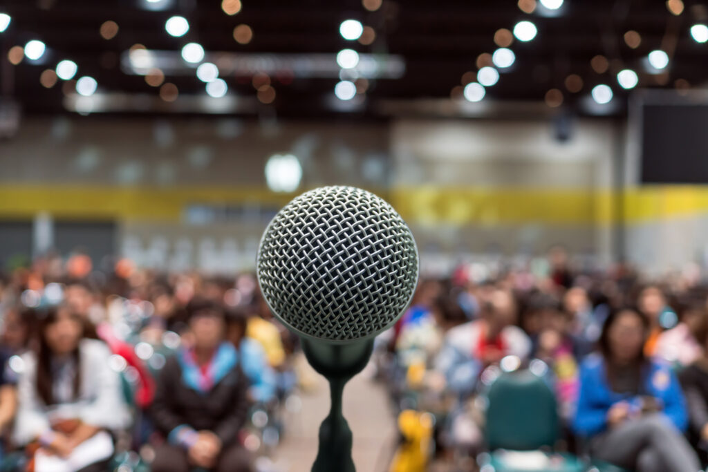 Microphone over the blurred photo of conference hall with attendees in the background; this image simulates a public speaker's perspective from behind a microphone.