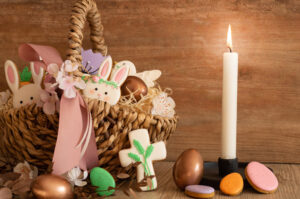 A photo of a lit, white, taper candle next to a basket full of Easter cookies shaped like bunnies and eggs and crosses