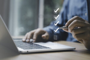 Man holding a pen in the air to check boxes with his left hand, while his right hand rests on a laptop keyboard.