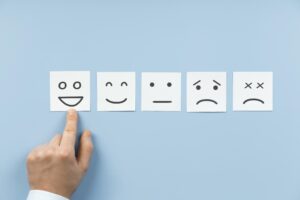 Five emoji faces on white square pieces of paper. A human finger points to the smiling face.